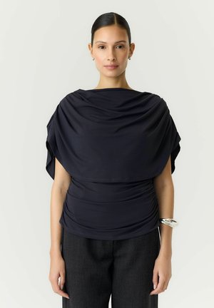 Femme aux cheveux raides attachés en arrière, portant un haut drapé foncé, un pantalon foncé, un bracelet en argent au poignet droit, debout devant un fond clair uni.