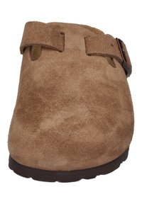 Bruine suède klomp met een gladde textuur, verstelbare band en een ronde neus. De zool is donker en plat, geschikt voor informele kleding.
