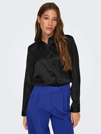 Schwarze Satin-Hemdbluse mit langen Ärmeln, kombiniert mit hochtaillierten blauen Hosen mit Falten und einer strukturierten Passform.