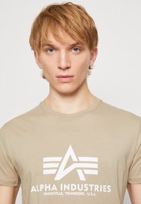 Tricou din bumbac bej cu guler rotund și mâneci scurte. Prezintă un design cu logo alb în centru, cu text dedesubt. Textură netedă.