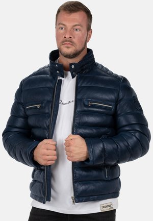 Mann, der eine marineblaue gesteppte Lederjacke auszieht, mit weißem T-Shirt und schwarzen Hosen, schaut leicht nach links vor einfarbigem Hintergrund.