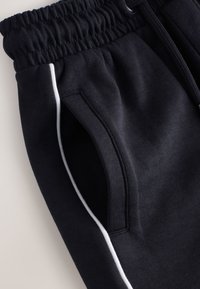 Close-up of black sweatpants pocket with white piping and elastic waistband with drawstring.