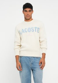 Bărbat îmbrăcat într-un pulover crem cu textul "LACOSTE Paris" și blugi de culoare bleu deschis, stând pe un fundal alb.
