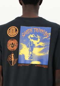 Tricou din bumbac negru cu un grafic mare albastru și galben, purtând textul "EARTH TRIPPING" și trei logo-uri portocalii pe partea stângă.