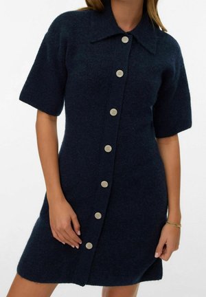 Žena oblečená v tmavě modrých vlněných šatech s krátkými rukávy, na knoflíky a s límečkem, s náramkem ze zlata na pravém zápěstí.