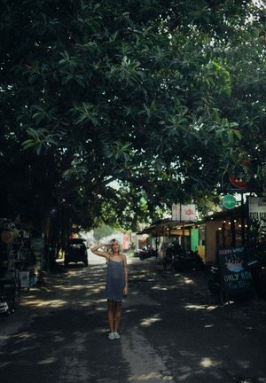 Szara sukienka bez rękawów, płaskie buty; postać stoi pod gęstym zielonym zadaszeniem drzew na ulicy z sklepami i pojazdami w tle.