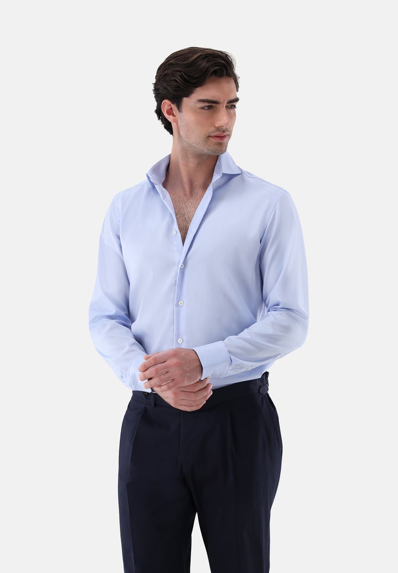Camicia azzurro chiaro con colletto alla francese, chiusura con bottoni e maniche lunghe, realizzata in tessuto liscio. Indossata con pantaloni scuri.