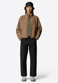 Brown lightweight jacket with high collar, green shirt underneath, black pants, and grey sneakers, paired with a black bucket hat.