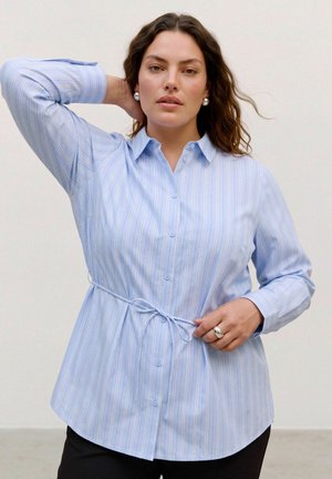 Le mannequin pose en chemise bleu clair rayée avec une ceinture et en pantalon noir. Elle a une main derrière la tête et tient la ceinture de l’autre.