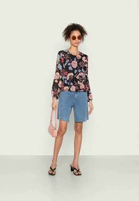 Blusa floral de mangas longas em preto com flores rosa e azuis, combinada com calções de ganga, uma bolsa rosa e sandálias de salto pretas.