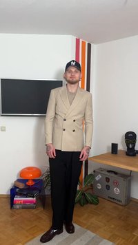 Beeži topeltbreasted pintsaku, hele kampsuni, mustade pükste ja pruunide kingadega. Seadistuses on teler, oranž lamp ja puidust laud.