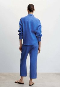 Blauw linnen overhemd en bijpassende cropped broek, met een losse pasvorm, lange mouwen en een rechte pijp. Draagt bij platte sandalen.