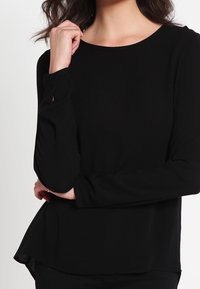 Haut noir à manches longues en tissu doux, avec un col rond, des poignets boutonnés et un ourlet légèrement évasé à l'arrière.