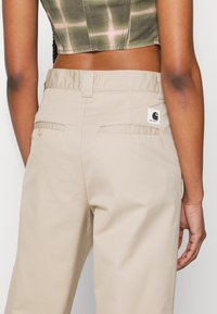 Pantalon en coton beige avec une coupe décontractée, doté de deux poches arrière et d'une petite étiquette de marque sur la ceinture.