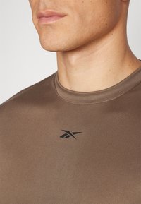 Brun athletic t-skjorte med rund hals, korte ermer og en svart logo på brystet. Glatt tekstur og tettsittende design.