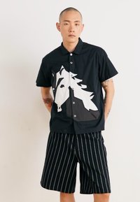 Black short-sleeve shirt with a white horse graphic, worn with black striped shorts. Features a classic collar and button closures.