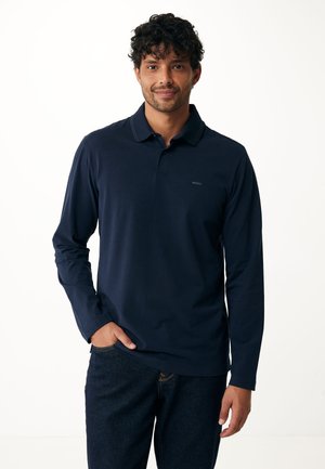 Camisa polo de manga larga en color navy con cuello, que presenta un logo sutil y un tejido suave y texturizado; combinada con jeans oscuros.