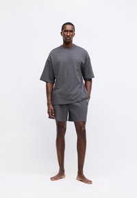 Tricou gri din bumbac cu mâneci scurte și largi și pantaloni scurți asortați. Design simplu cu o croială relaxată, prezentat pe un fundal neutru.