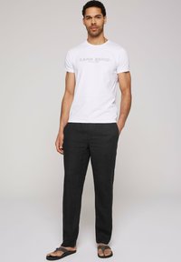 Witte katoenen t-shirt met "CAMP DAVID" in grijze tekst, gecombineerd met donkere linnenbroeken en slippers. Eenvoudig ontwerp en losse pasvorm.