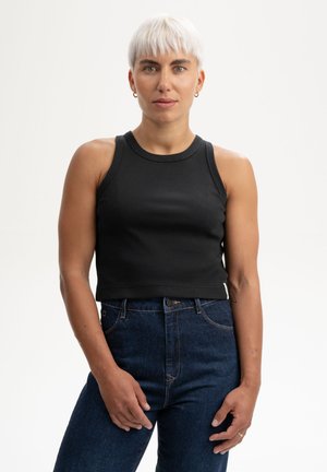 Jonge persoon met kort platina haar, gekleed in een zwart mouwloos topje en donkerblauwe spijkerbroek, staand tegen een witte achtergrond.