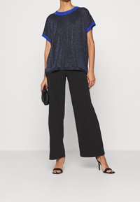 Granatowa bluzka z metalicznym połyskiem, okrągły dekolt i niebieskie akcenty. W połączeniu z czarnymi spodniami w szerokiej nogawce i sandałami na obcasie.