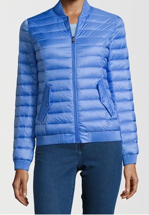 Femme portant une veste matelassée bleu clair zippée avec col, poignets, ourlet côtelés et deux poches avant à rabat, associée à un jean bleu foncé.