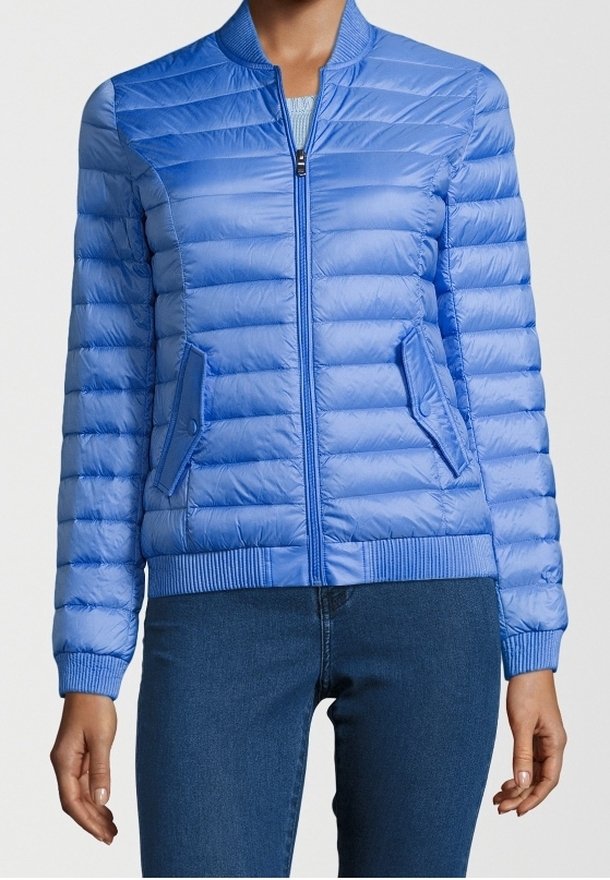 Femme portant une veste matelassée bleu clair zippée avec col, poignets, ourlet côtelés et deux poches avant à rabat, associée à un jean bleu foncé.