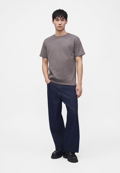 Jeune homme portant un t-shirt gris à manches courtes, un jean bleu foncé à jambes larges et des chaussures noires, debout avec une main dans la poche.