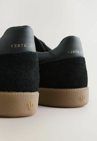 Zapatillas negras con un empeine de ante y cuero, suela de goma y texto en relieve dorado. Presenta un logo distintivo de corona en el talón.
