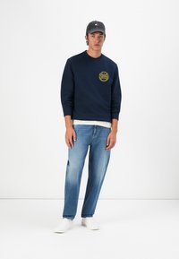 Sudadera marine con logotipo amarillo, combinada con jeans de color azul claro y zapatillas blancas. El modelo lleva una gorra gris y una camiseta blanca debajo.