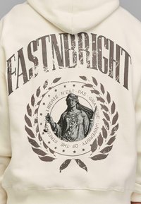 Krémová mikina s kapucňou, na ktorej je vytlačený text "FASTNBRIGHT" veľkými, ošúchanými hnedými písmenami a grafika dámy s váhami obklopenú vavrínovými listami.