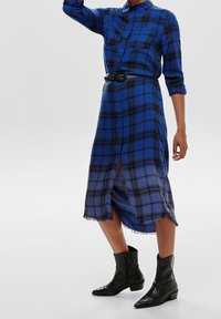 Niebieska sukienka w kratę z długimi rękawami, zapinana na guziki, z postrzępionym dołem. Noszona z czarnym paskiem i botkami za kostkę.
