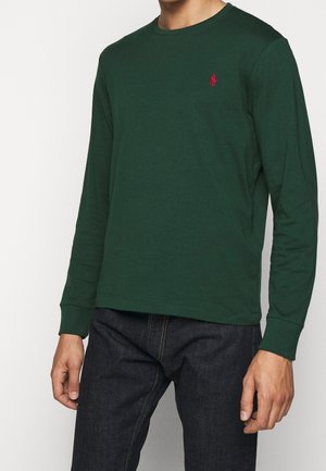 Mann trägt ein dunkelgrünes Langarmshirt mit rotem Logo auf der Brust und dunkle Jeans, steht vor neutralem Hintergrund.