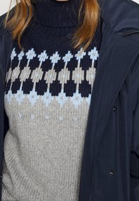 Pull en tricot avec un col montant et des motifs géométriques en bleu marine, bleu clair et beige sur un fond gris. Porté sous une veste extérieure foncée.