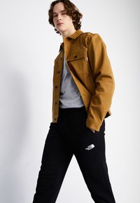 Bruine, kraagloze jas over een grijs sweatshirt, gecombineerd met zwarte joggingbroeken met een wit logo. Glad textuur met knoopdetails.