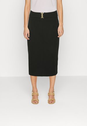 Kvinde iført en sort knælang pencilskirt med en guldlåsdetalje, hvid top og beige åbne tå-hælsandaler, stående på en hvid gulv.