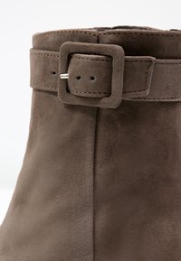 Bottine en daim marron avec une sangle à boucle carrée, texture douce et lignes épurées, mettant en avant un design minimaliste.