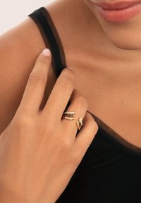 Guldringar med klara och färgade stenar, med en vriden design. En hand vilar försiktigt på en axel med ett svart nätband.