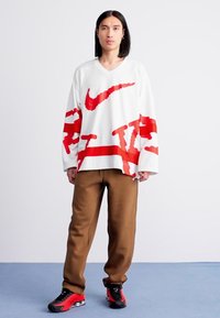 Mand med langt sort hår iført en hvid oversized sweater med røde grafik, brune bukser og røde-sorte sneakers, der står på et blåt gulv.