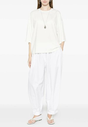Donna che indossa pantaloni bianchi larghi, top bianco oversize, sandali infradito bianchi e una collana con ciondolo argento su uno sfondo neutro.