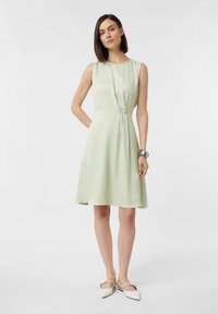 Hellgrünes, ärmelloses Kleid mit rundem Ausschnitt und gefälteltem Detail an der Taille, kombiniert mit weißen spitzen Schuhen. Glatte Textur.