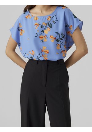 Vrouw draagt een losse lichtblauwe blouse met oranje bloemenprint, ingestopt in een zwarte broek met hoge taille tegen een effen achtergrond.