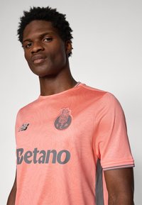Camiseta de fútbol rosa con un patrón geométrico, acentos grises, cuello redondo y logotipos de New Balance y Betano en el frente.