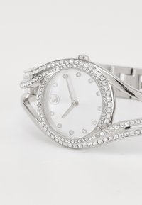 Orologio argento con quadrante rotondo, decorato con cristalli trasparenti su un bracciale ondulato. Il quadrante bianco presenta indicatori e lancette minimaliste.