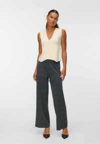 Débardeur sans manches en crème avec décolleté en V et fermeture à boutons, accompagné de pantalons à carreaux bleu et noir à jambe large, accentués par des chaussures pointues marron.