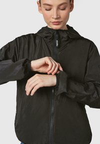 Crna lagana jakna s kapuljačom, prednjim zipom, elastičnim manžetama i minimalističkim dizajnom. Materijal izgleda glatko i otporan na vodu.