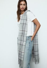 Écharpe à carreaux gris en matériau doux, avec des bords frangés et un motif à carreaux en gris clair et foncé, drapée sur une chemise blanche.