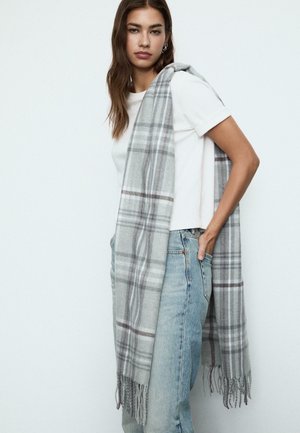 Écharpe à carreaux gris en matériau doux, avec des bords frangés et un motif à carreaux en gris clair et foncé, drapée sur une chemise blanche.