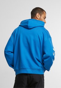 Felpa blu con cappuccio ampio, orlo a costine e maniche a sbuffo. Presenta una grafica bianca sulla manica sinistra. Tessuto morbido e strutturato.