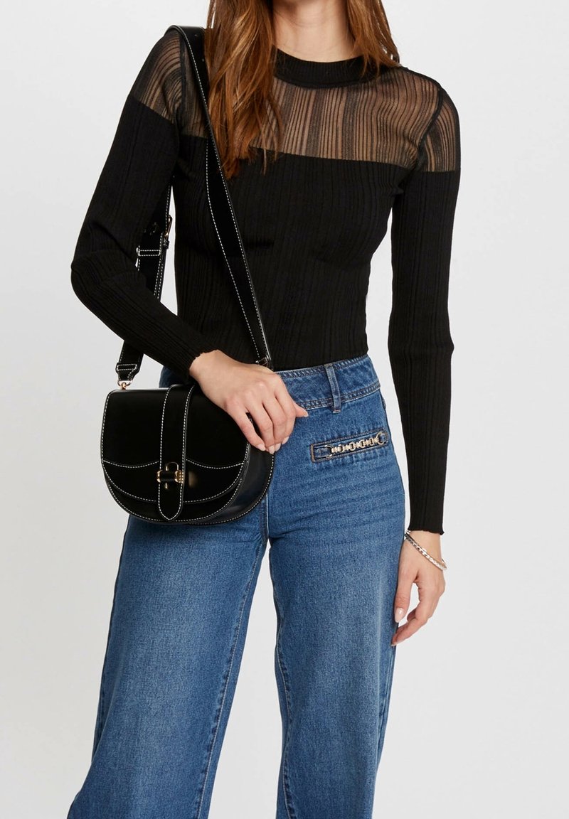 Femme portant un haut noir transparent à manches longues et un jean bleu, tenant un sac bandoulière noir avec des coutures blanches et une fermeture dorée.